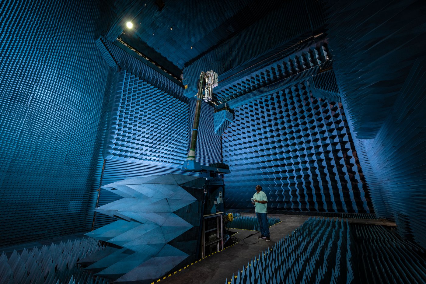 A spacesuit test article standing in an anechoic chamber whose walls and floor are covered with blue foam absorber pyramids, with an engineer beside it