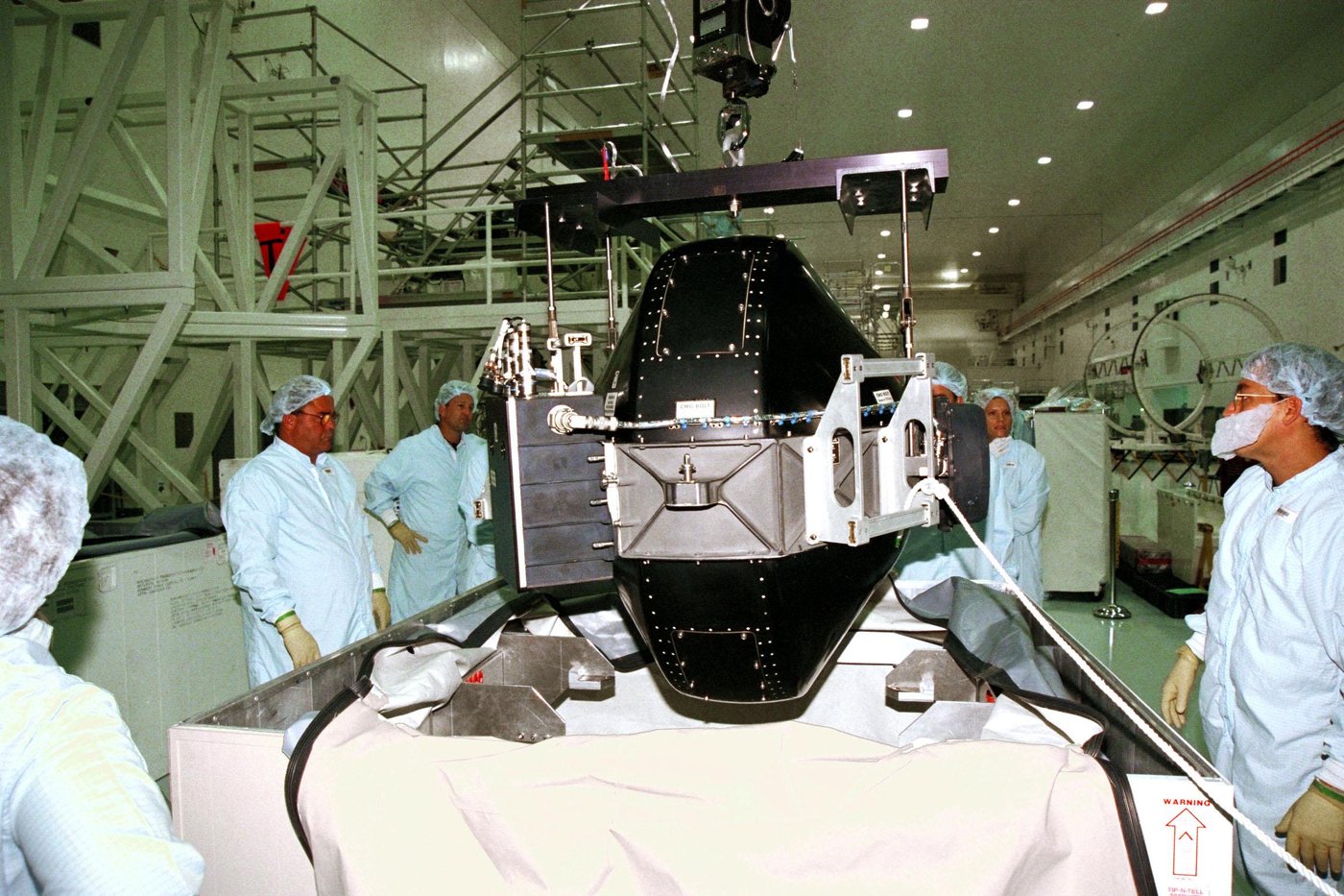 Technicians in a clean room at Kennedy Space Center lifting one of the International Space Station's control moment gyroscopes, a black housing on a crane, out of its shipping container